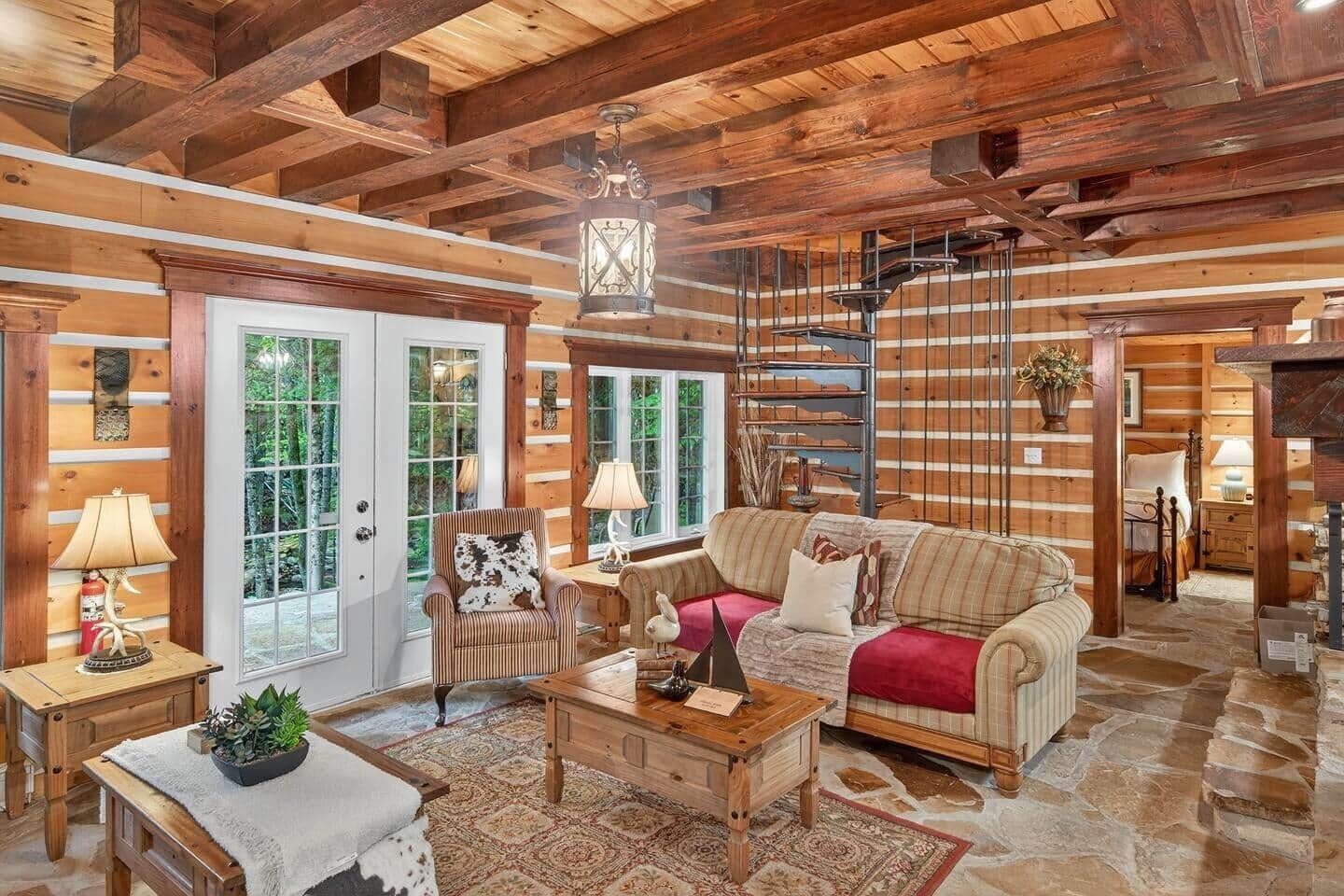 A log cabin interior with a wooden ceiling, a sofa, a chair, a table, and a lamp.