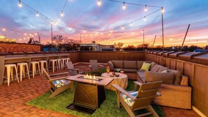 An outdoor patio area with a fire pit, seating, and string lights against a sunset sky.