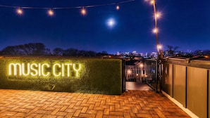 A nighttime view of a rooftop area with a neon sign reading "MUSIC CITY" and a brick pathway leading to a building entrance.