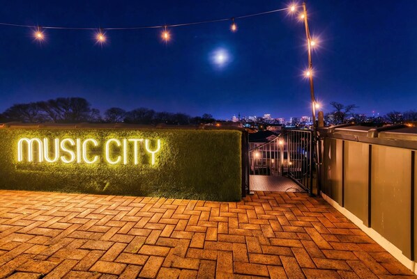 A nighttime view of a rooftop area with a neon sign reading "MUSIC CITY" and a brick pathway leading to a building entrance. - Luxury Retreat Double Rooftop Deck Game Room (Nashville)