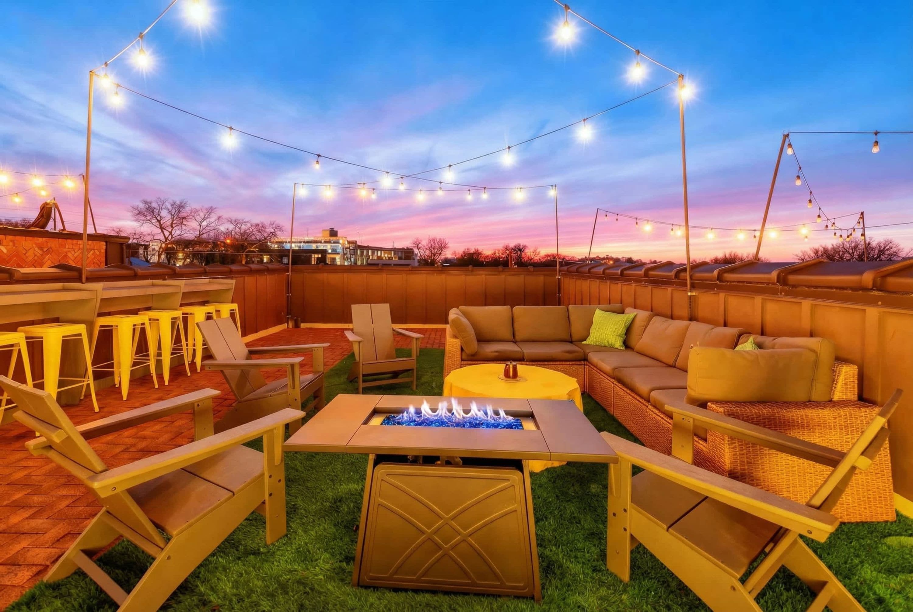 An outdoor patio with string lights, a fire pit, seating, and a view of the city skyline.