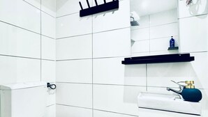 Une salle de bain avec des murs recouverts de carreaux blancs, des toilettes, un lavabo avec un distributeur de savon bleu et un miroir avec une étagère au-dessus.