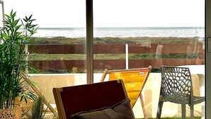 Ein Zimmer mit großem Fenster und Blick auf einen Strand. Ein Sessel mit Kissen, ein kleiner Tisch und zwei Kübelpflanzen sind vorhanden.