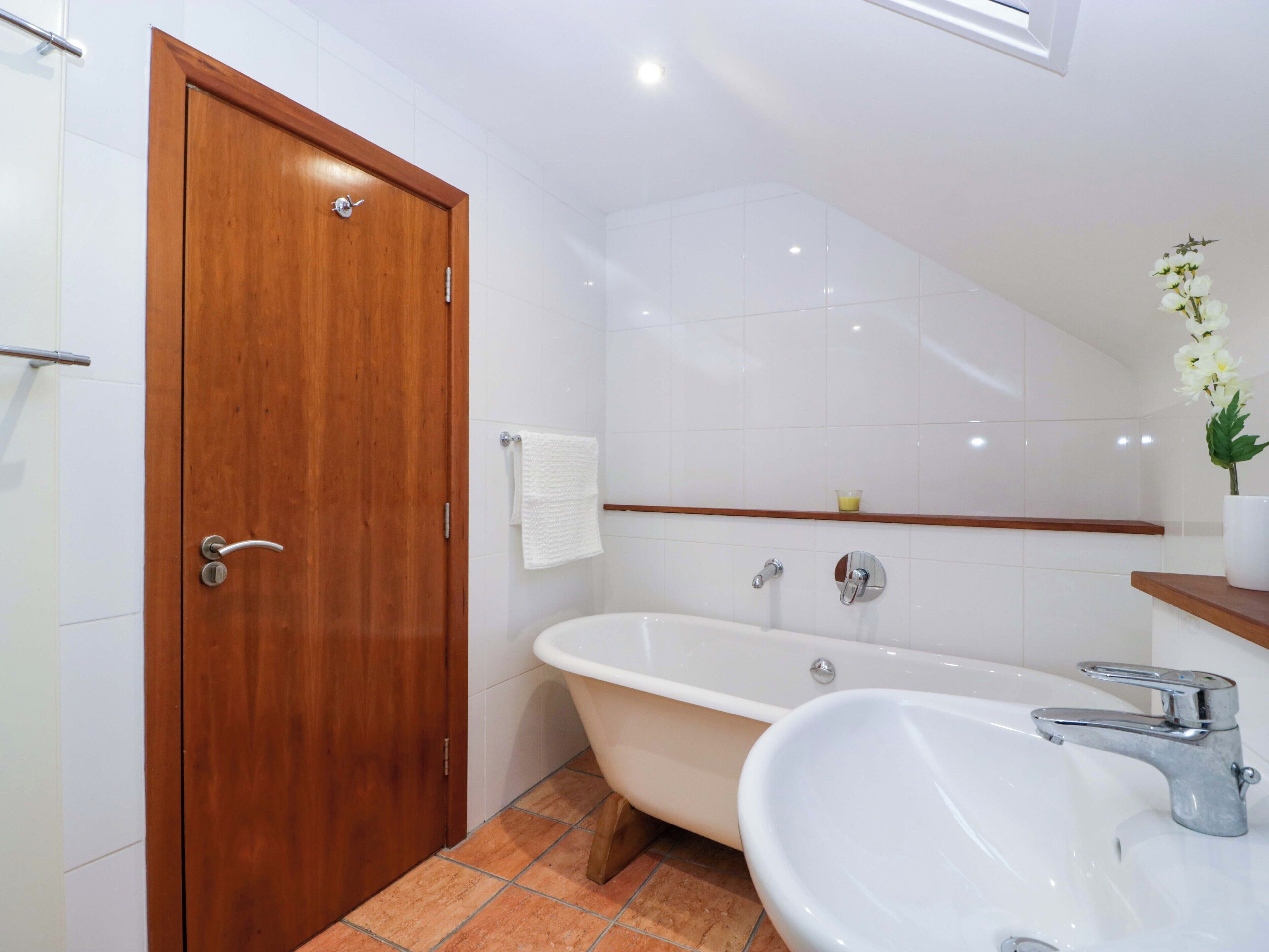 A bathroom with a wooden door, white tiles, a bathtub, a sink, and a small shelf with a plant.
