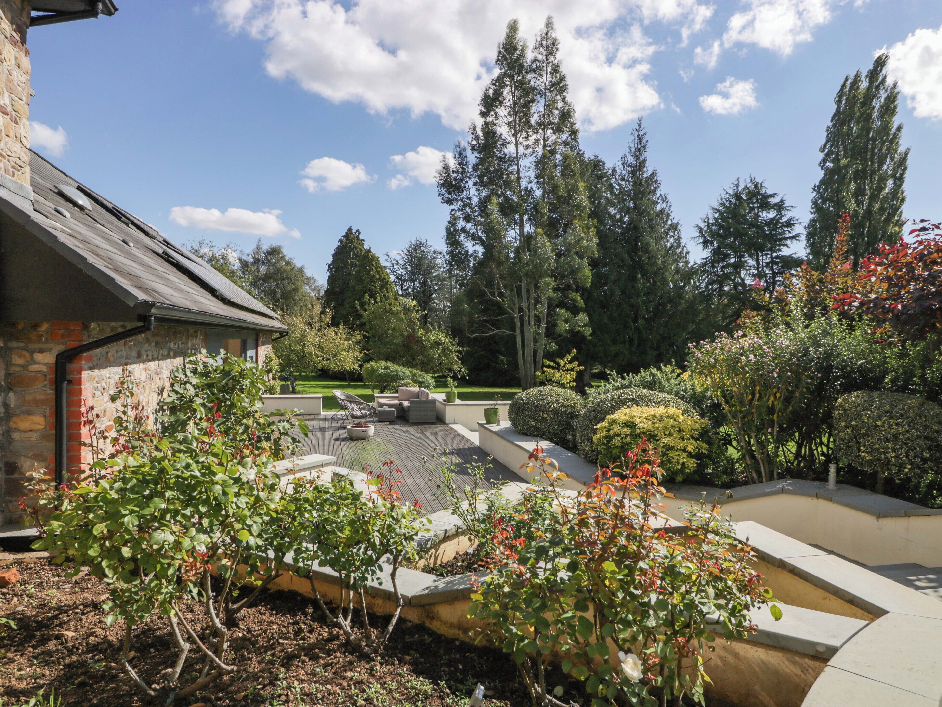 A landscaped garden with a patio area, a stone building, and tall trees.