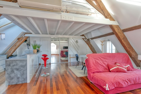 Ein Loft-Zimmer mit Holzbalkendecke, einem roten Sofa, einer Küchenzeile und einem Esstisch.