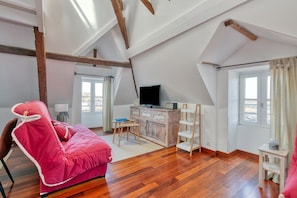 Ein Zimmer mit Holzboden, einem roten Sessel, einem Fernseher, einem Holzschrank und einem Fenster mit Vorhängen.