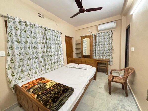 A bedroom with a bed, a wooden chair, a desk, a mirror, and a ceiling fan. - Sri Mayapur Chandrodaya Guest House (Krishnanagar)