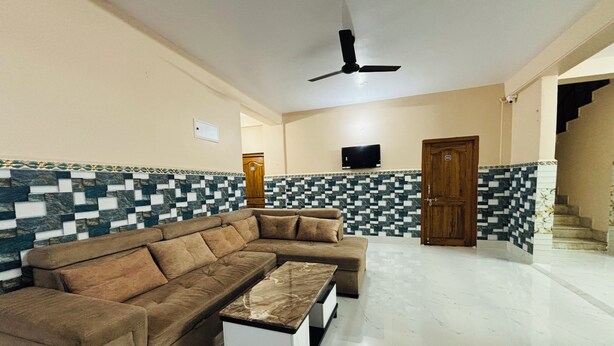 A spacious living area with a brown sectional sofa, a small white side table, a ceiling fan, a television mounted on the wall, and a patterned tiled wall. - Sri Mayapur Chandrodaya Guest House (Krishnanagar)