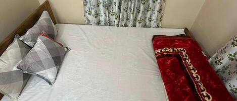 A bed with a white sheet, a red blanket with gold trim, and two pillows, one with a geometric pattern and another with a floral design.