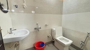A bathroom with a sink, toilet, shower, and a red bucket. - Sri Mayapur Chandrodaya Guest House (Krishnanagar)