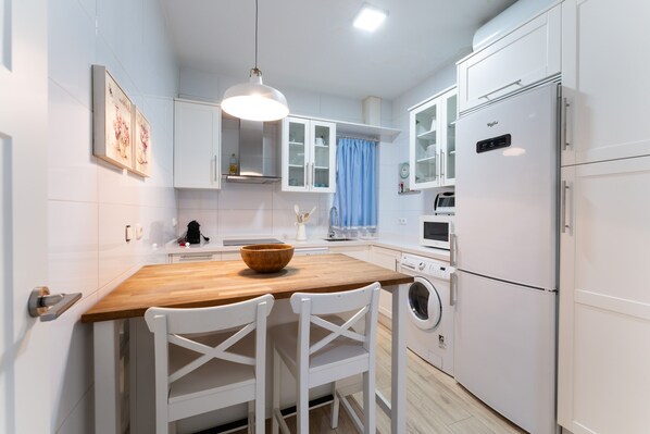 Une cuisine moderne avec des meubles blancs, un îlot central en bois, des chaises blanches, un lave-linge et un réfrigérateur.