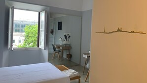 Ein Schlafzimmer mit einem Bett, einem Schreibtisch und einem Sessel. Ein Fenster bietet Blick auf Gebäude und Bäume.