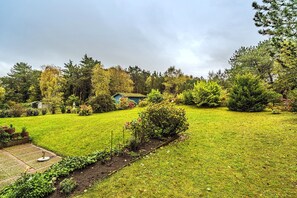 A well-maintained garden with a variety of plants and trees, a small house, and a clear sky.