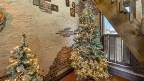 A spacious room with a high ceiling and exposed brick walls. There are two large Christmas trees with lights, and a staircase with wooden railings.