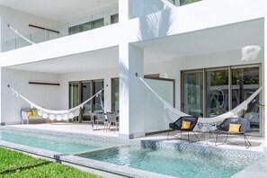 Un área de piscina moderna al aire libre con una hamaca, asientos y un porche con paredes de vidrio.