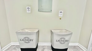 Dos cestos grises para ropa con bases negras y el texto "DELUXE LAUNDRY SERVICE" en el centro, ubicados sobre un piso de mármol en un pasillo con paredes de tonos claros.
