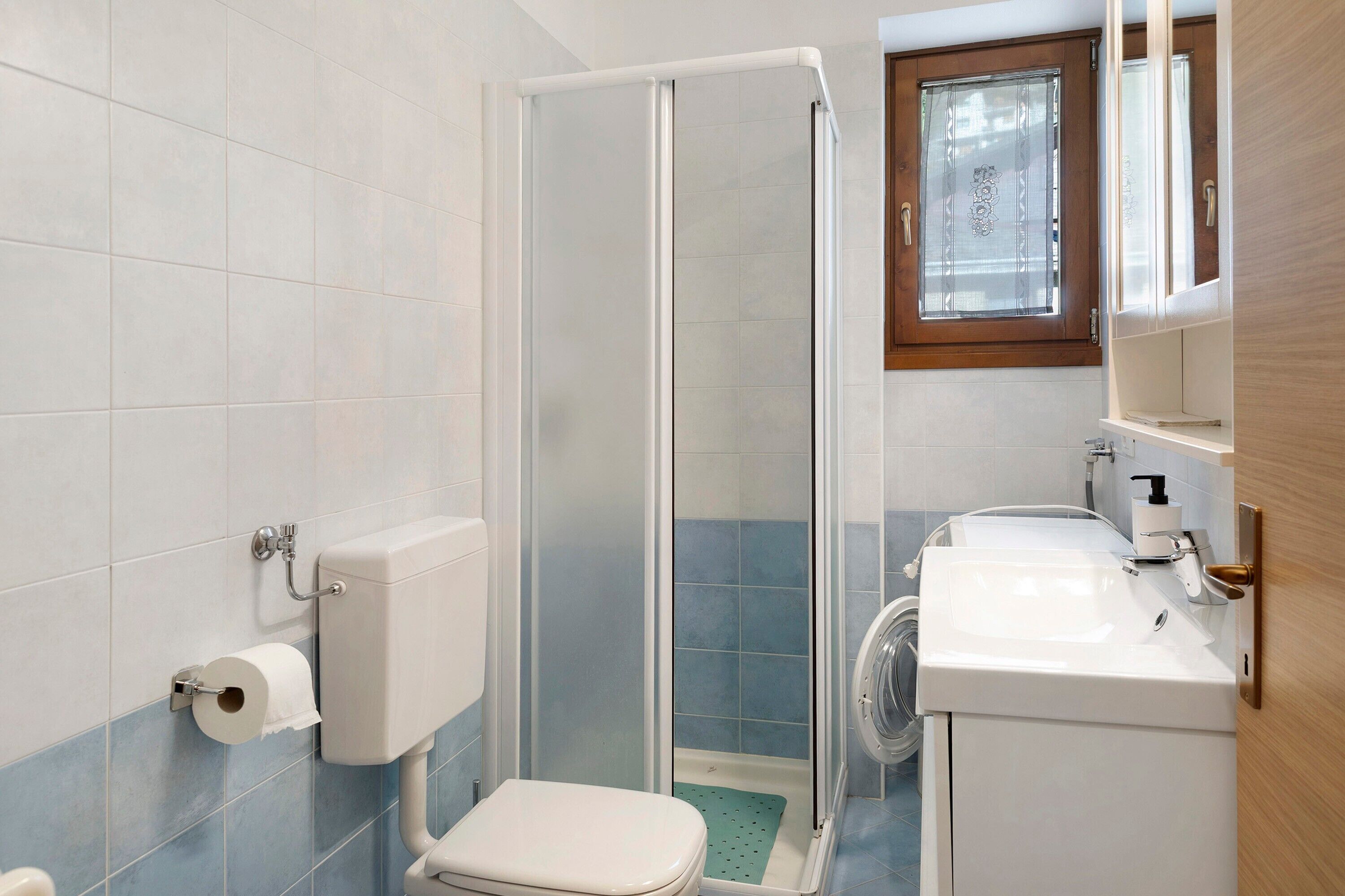 A bathroom with a toilet, shower, sink, and a window with a curtain.