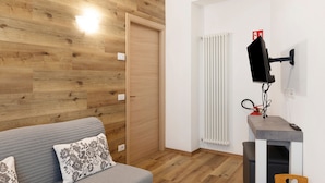 Quarto com chão de madeira, sofá cinza com almofadas estampadas, radiador branco e televisão na parede.