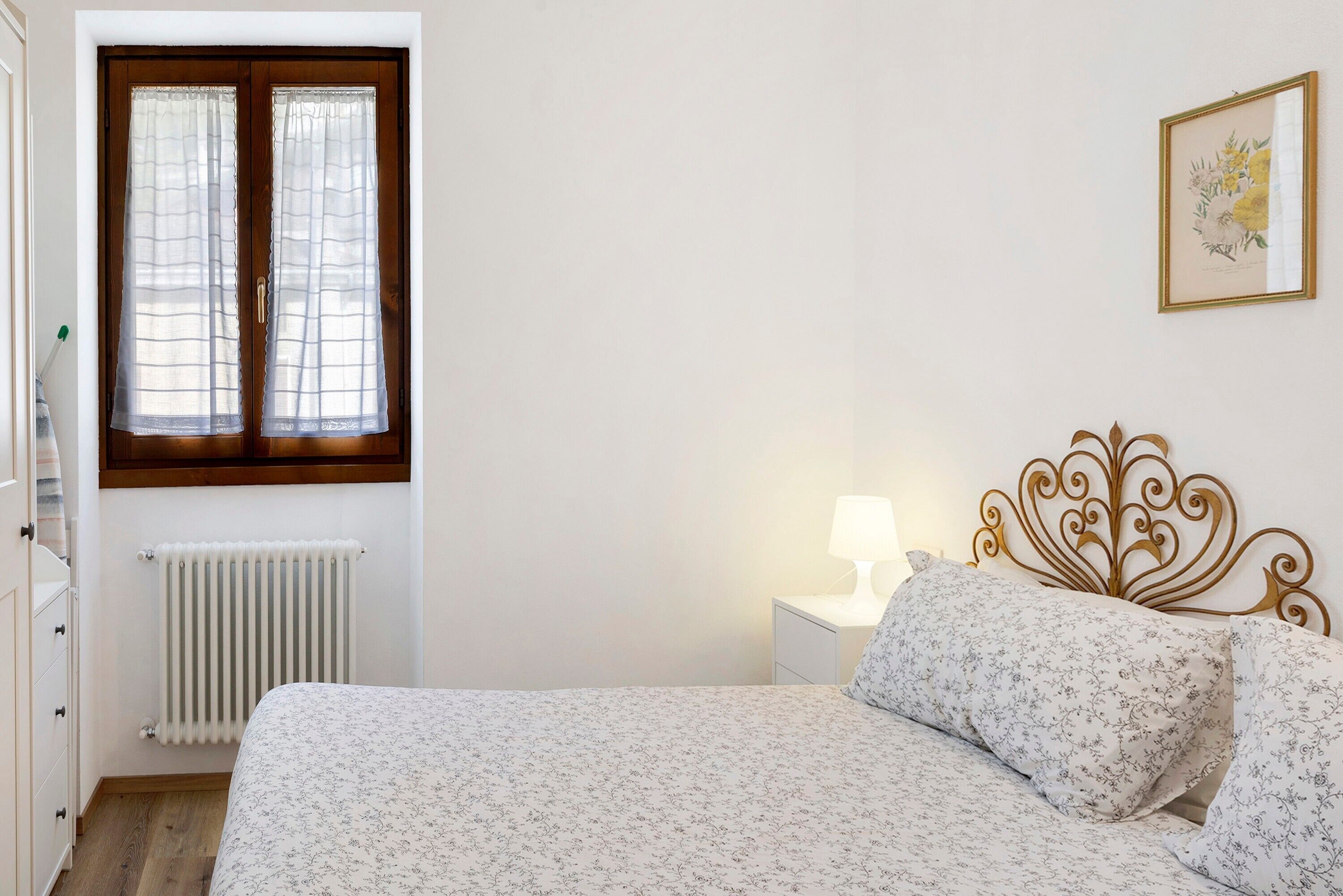 Une chambre à coucher avec un lit, un radiateur, une fenêtre avec des rideaux, une table de chevet avec une lampe et un tableau encadré au mur.