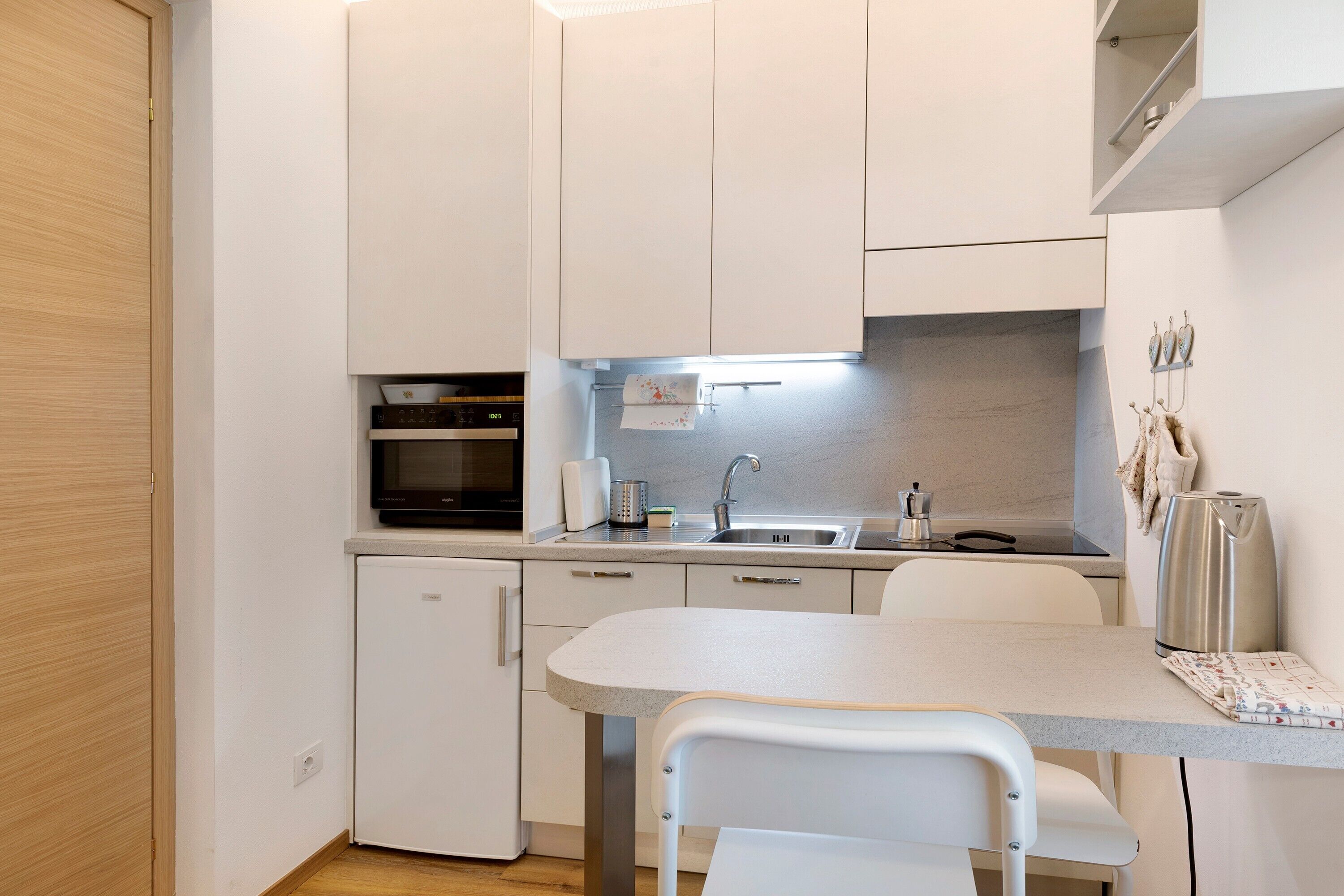 Une cuisine compacte avec des meubles blancs, un four encastré, un évier et un petit réfrigérateur.