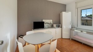 Een kleine, moderne kamer met een witte tafel en stoelen, een flatscreen televisie, een magnetron en een raam met uitzicht op gebouwen.