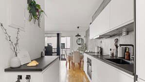 Una cocina moderna con armarios blancos, encimera negra y suelo de madera. Hay un área de comedor con mesa y sillas, y vistas al exterior a través de un amplio ventanal.