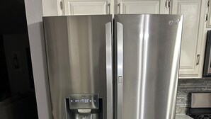Een koelkast van roestvrij staal met een waterdispenser, witte keukenkastjes en een betegelde achterwand in een keuken.