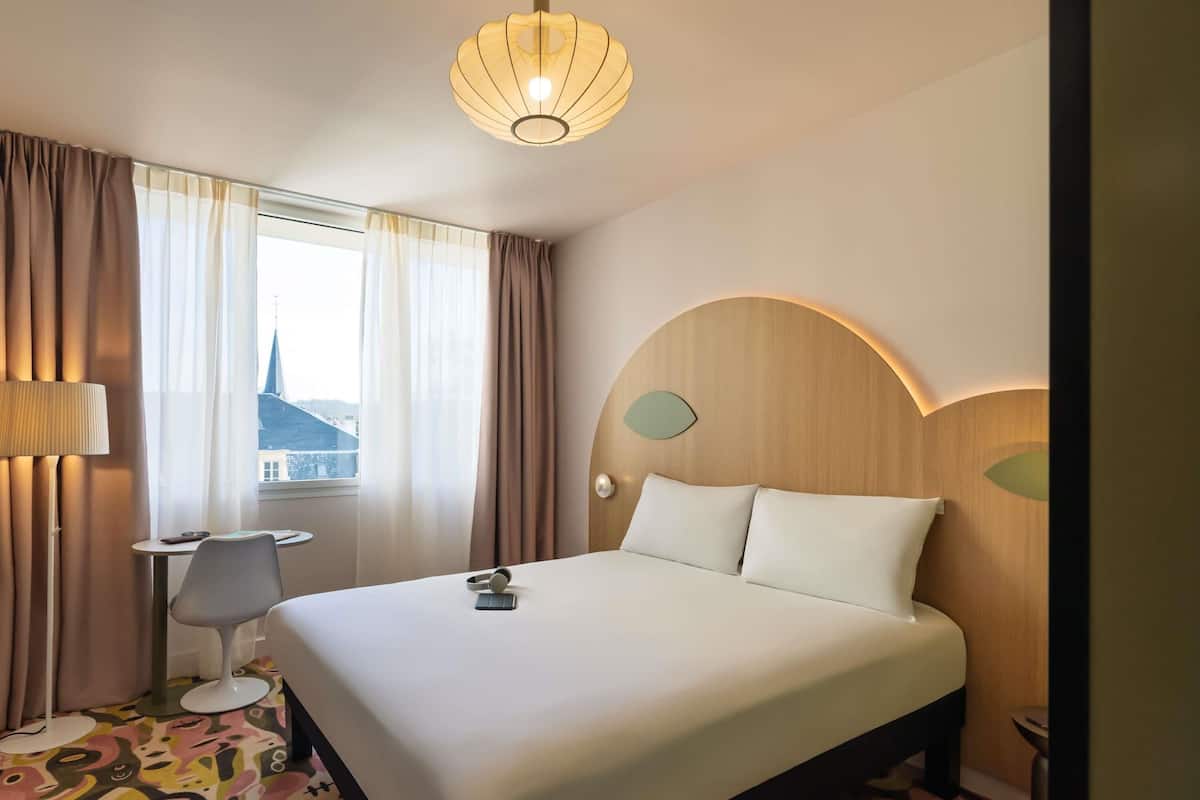 A hotel room with a large bed, a desk with a chair, a lamp, and a view of a building outside.