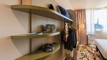 Quarto de hotel com cama, escrivaninha, cadeira, janela com vista e prateleiras com roupas e uma mala.