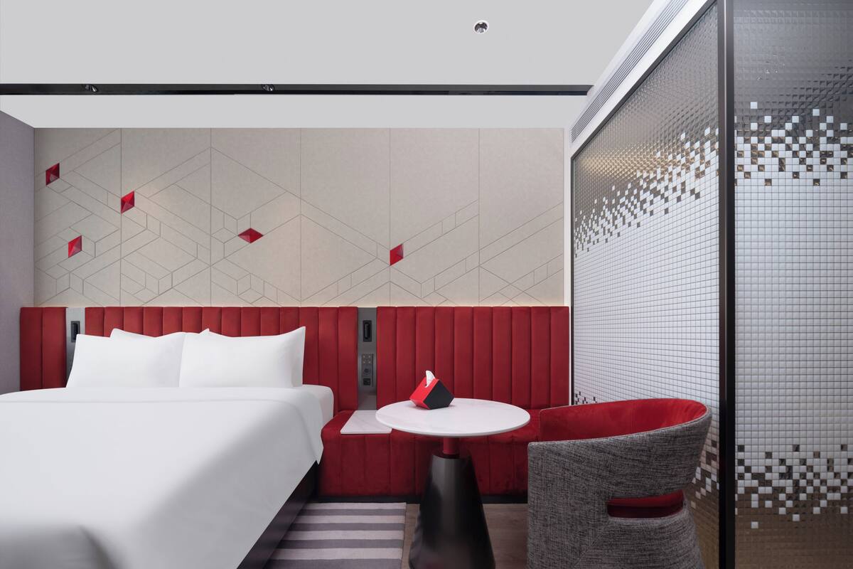 Une chambre d’hôtel moderne, équipée d’un lit, d’une petite table, d’un fauteuil rouge recouvert de tissu et d’une cloison en verre.