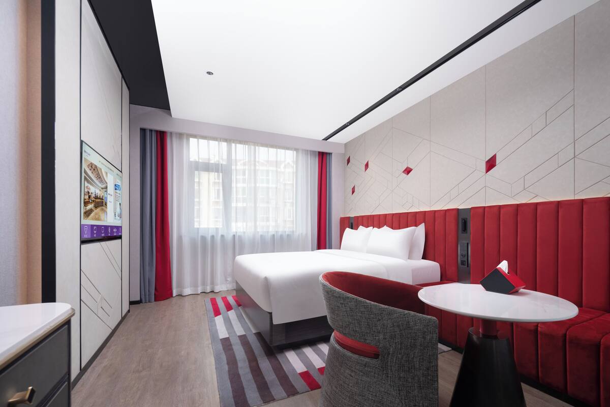 Une chambre d’hôtel moderne avec un lit, un canapé rouge recouvert de tissu, une table ronde et une grande fenêtre avec des rideaux.