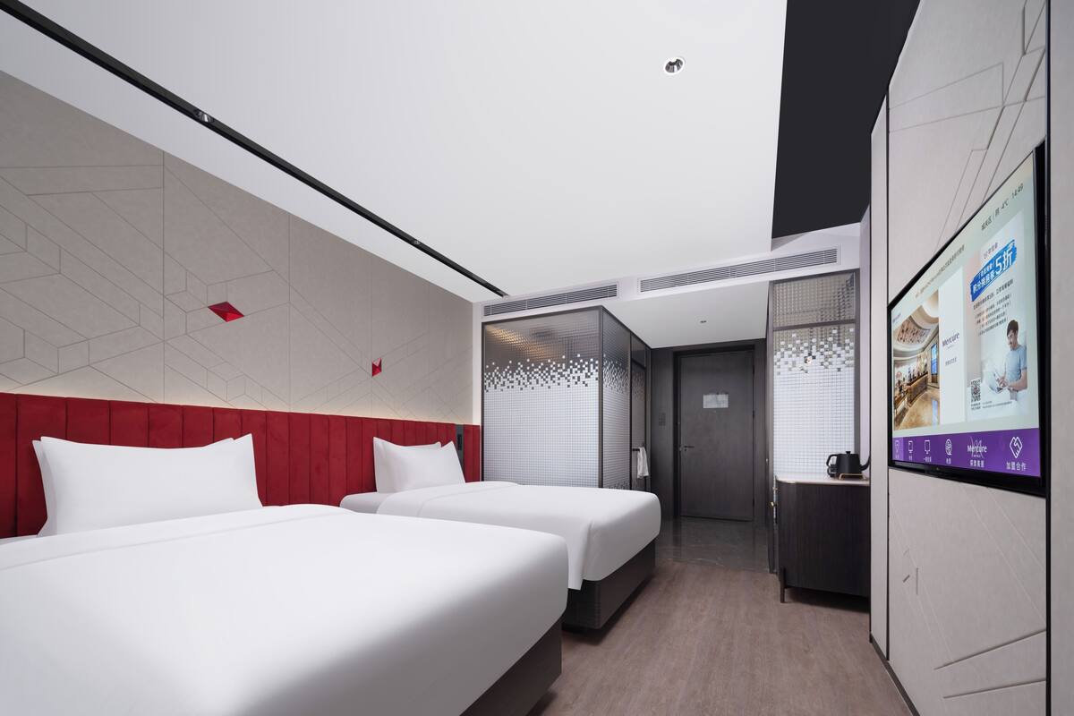 Une chambre d’hôtel avec deux lits, une tête de lit rouge, un grand écran et un mur recouvert de carreaux.