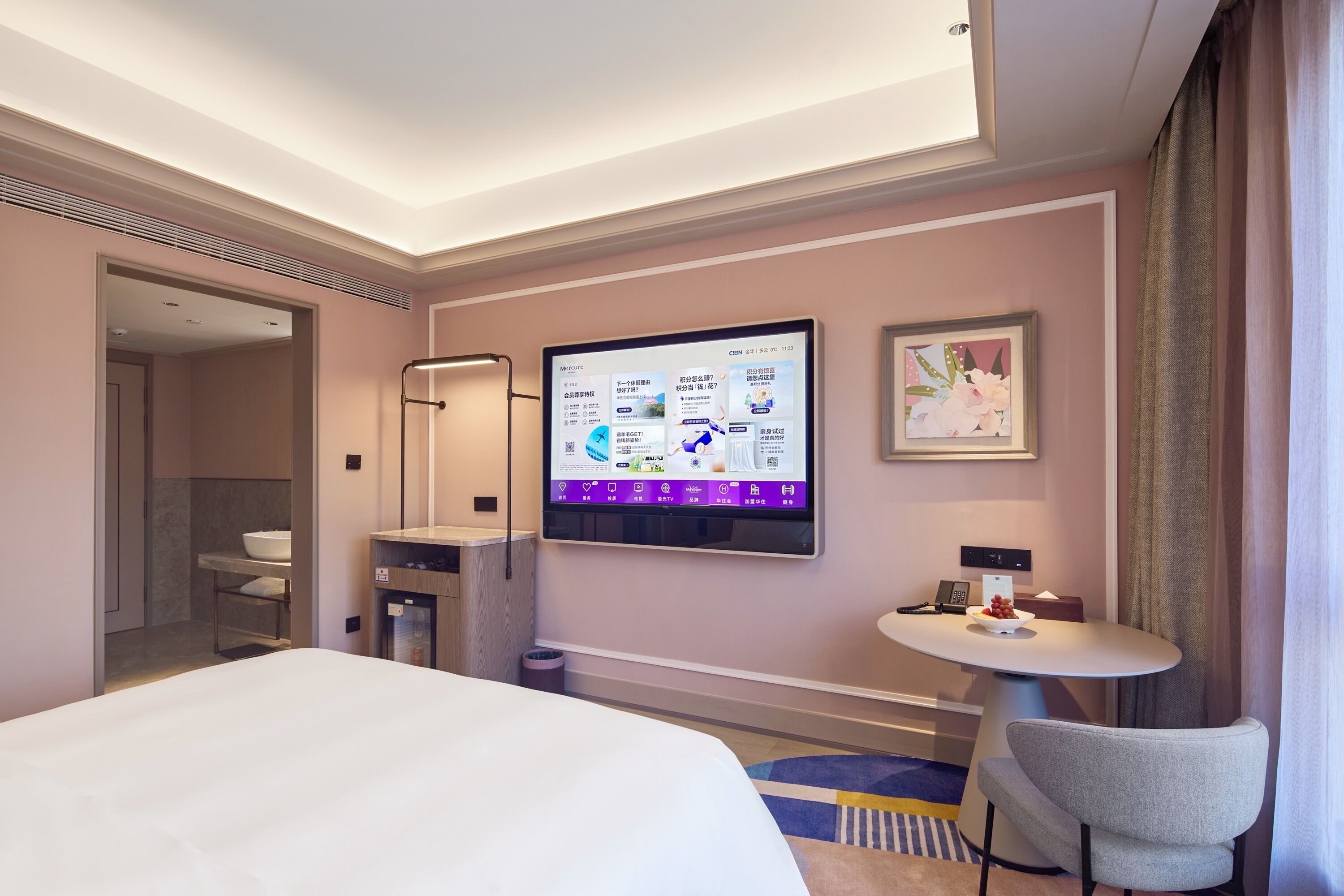 A hotel room with a bed, a TV mounted on the wall, a small table with a chair, and a bathroom visible through an open door.