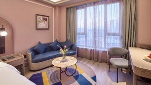 Hotelkamer met een bank, een eettafel, een stoel, een klein bijzettafeltje met bloemen, een bed, een nachtkastje en een groot raam met gordijnen.