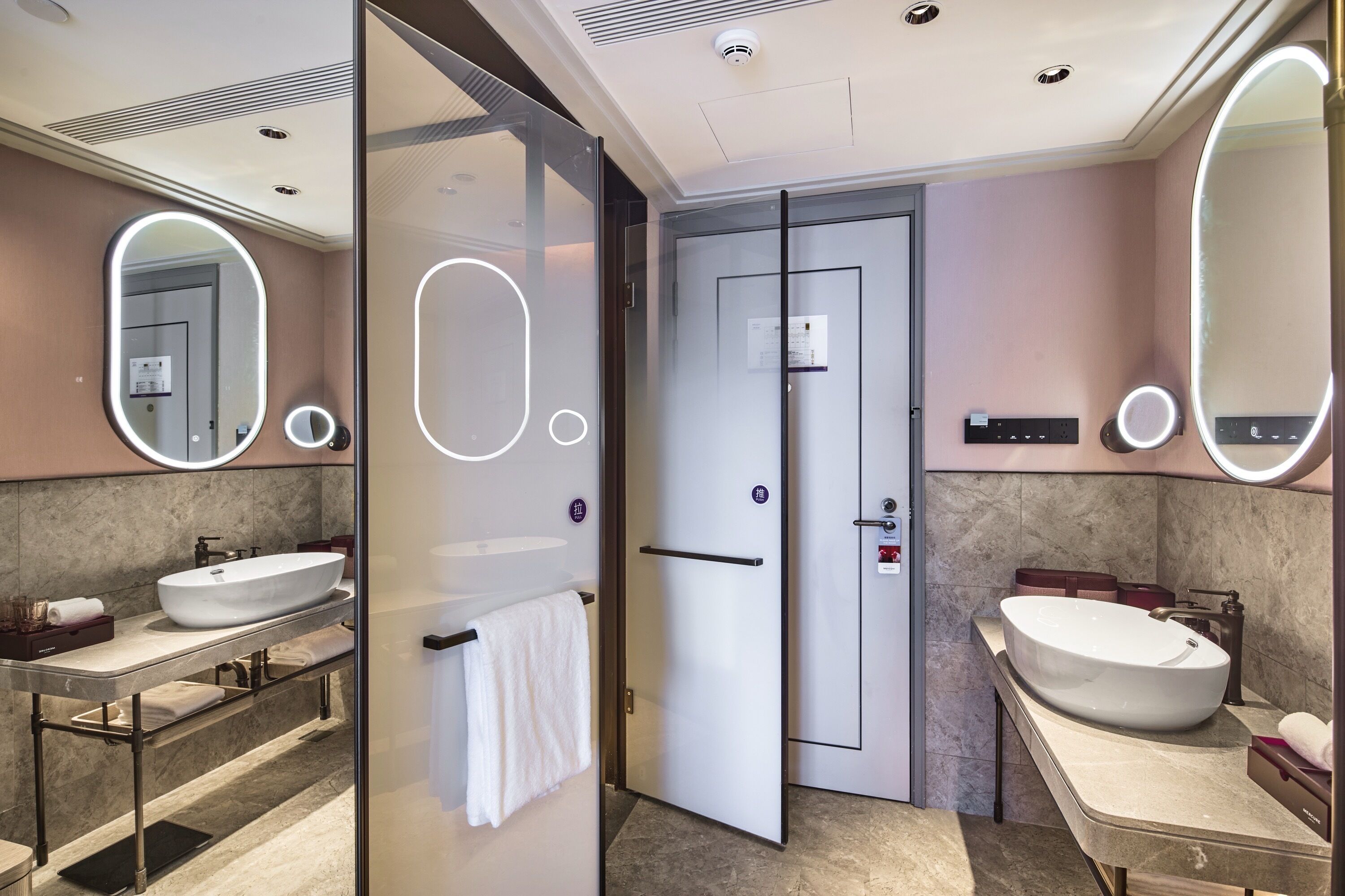 Un baño moderno con dos lavamanos, mampara de vidrio y puerta con panel de control.