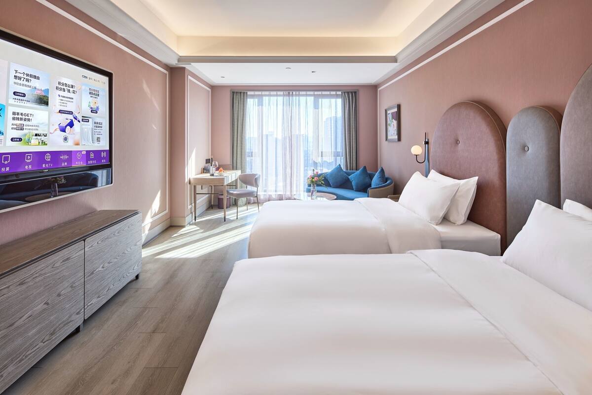 Ett hotellrum med två sängar, en tv som är monterad på väggen, ett skrivbord med en stol och en soffa med blå kuddar.