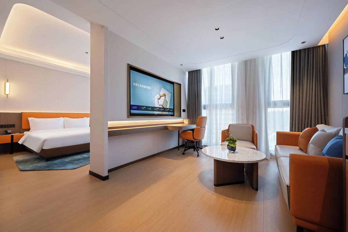Une chambre d’hôtel moderne équipée d’un lit, d’un téléviseur fixé au mur, d’un coin salon avec une table basse et de grandes fenêtres avec des rideaux.