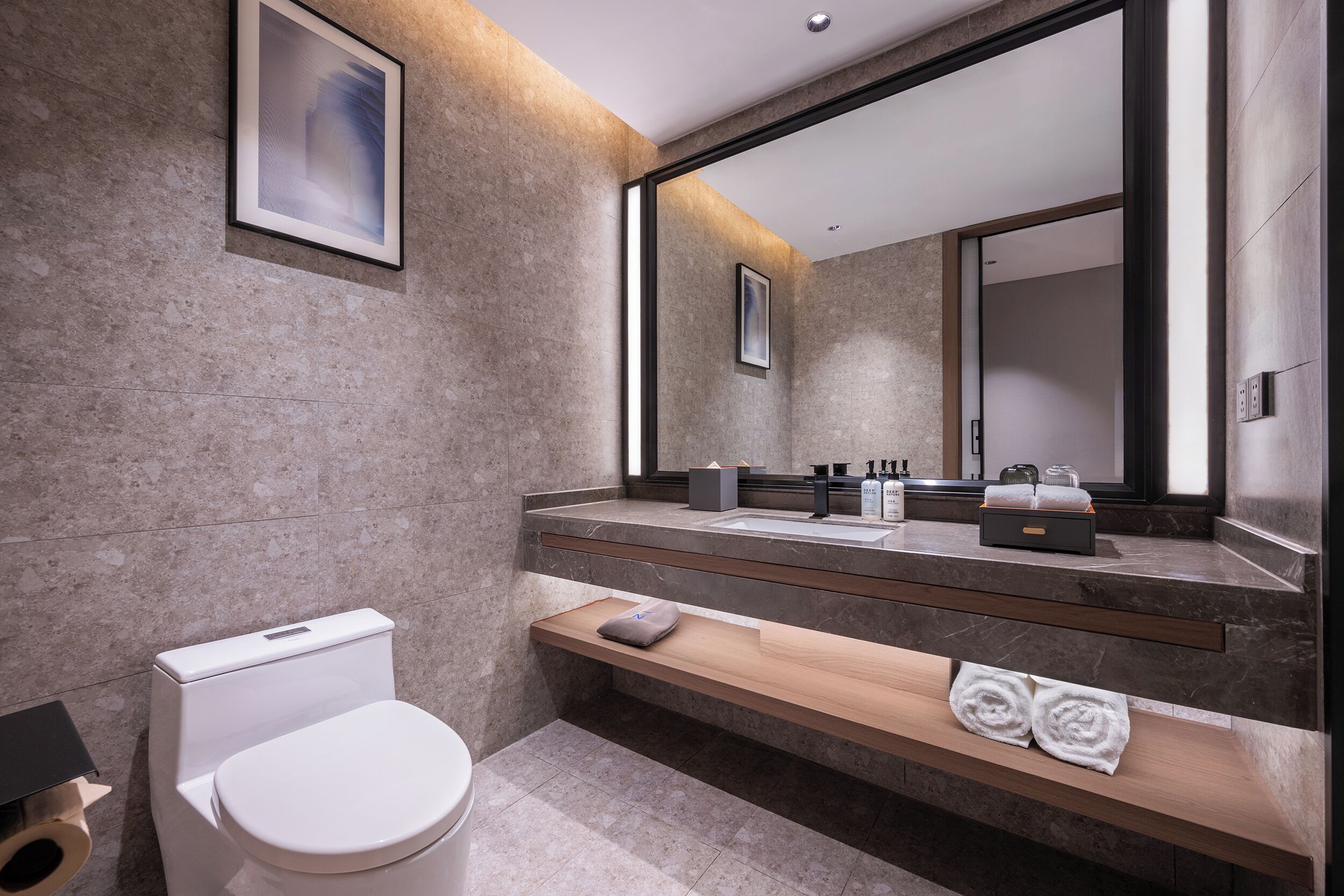 Ein modernes Badezimmer mit einer weißen Toilette, einem Holzwaschtisch, einem großen Spiegel und einer Marmorwand.