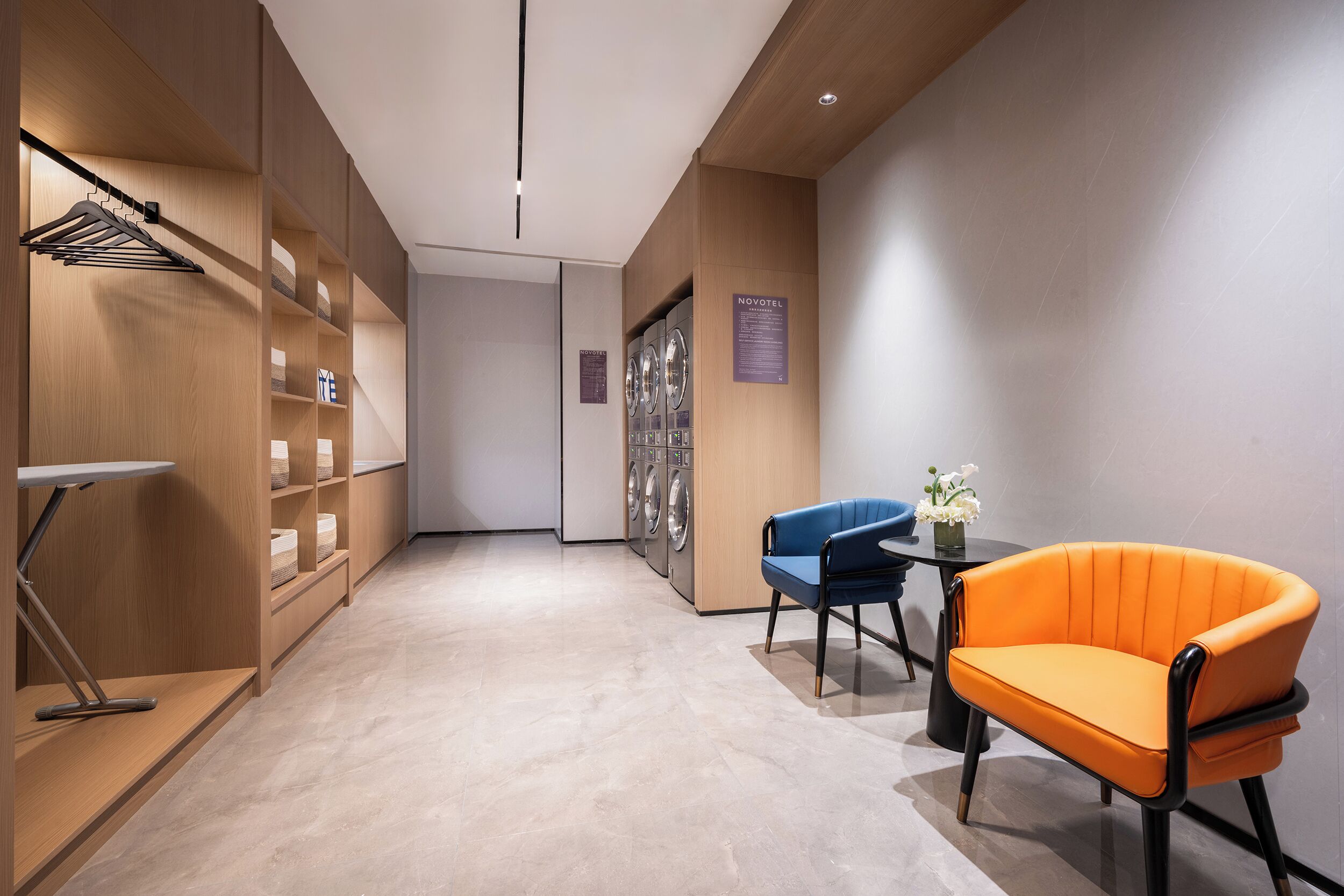 A hotel lobby with a wooden reception desk, a blue chair, an orange chair, and a wall-mounted laundry machine.