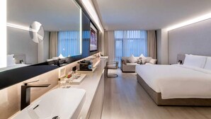 Ein modernes Hotelzimmer mit einem großen Bett, einer separaten Couch, einem großen Spiegel, einem Flachbildfernseher, einer Minibar und einem Badezimmer mit einem großen Waschbecken und Spiegel.
