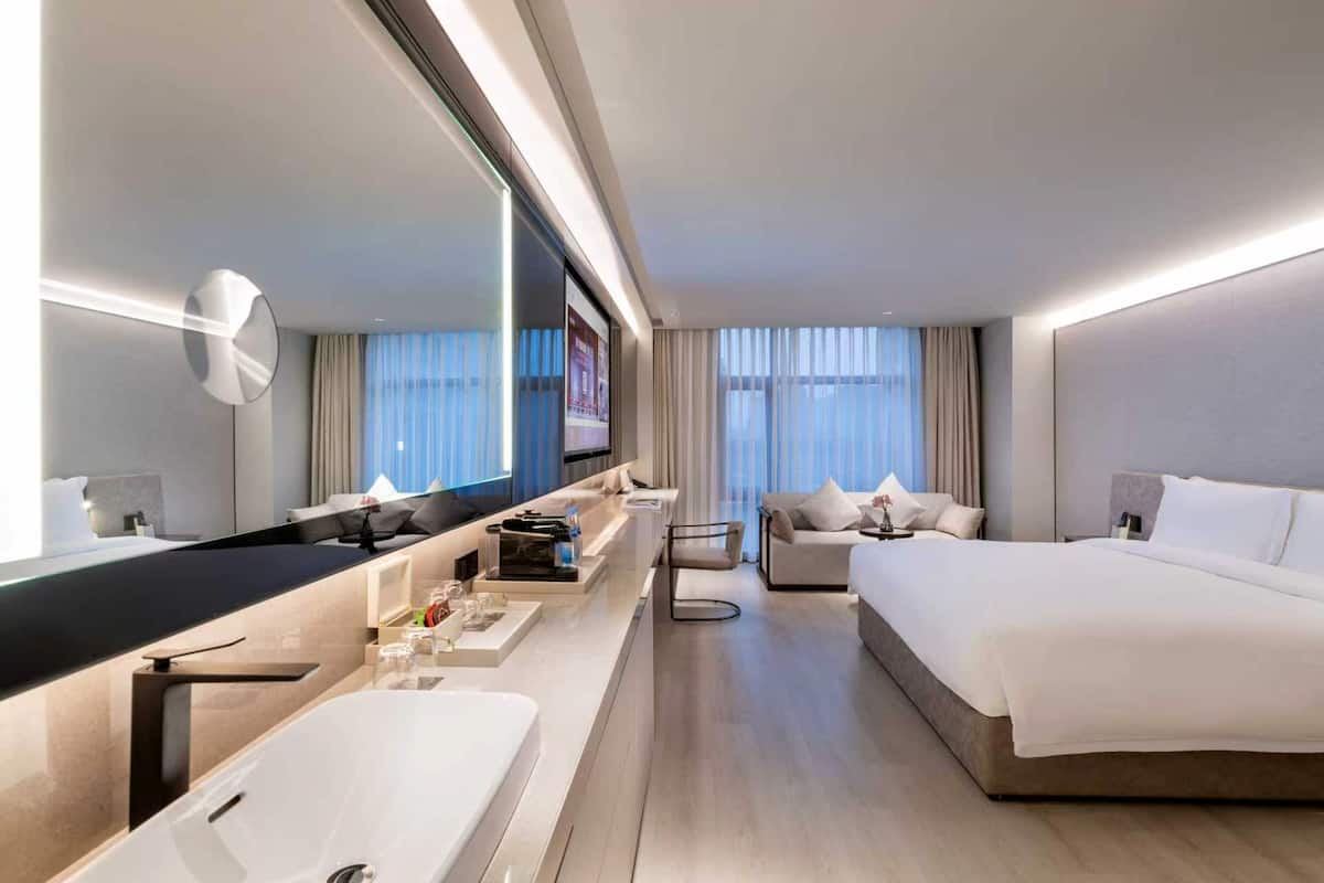 Een moderne hotelkamer met een groot bed, een aparte zithoek, een grote spiegel, een flatscreen televisie, een minibar, en een badkamer met een ruime wastafel en spiegel.