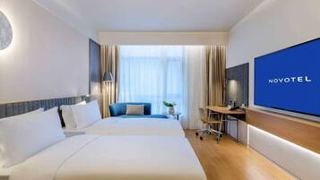 A hotel room with two beds, a desk, a TV, and a large window with curtains.