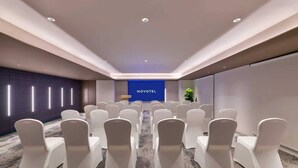 Ein Konferenzraum mit Reihen weißer Stühle, einem Rednerpult und einer blauen Wand mit der Aufschrift 'NOVOTEL'.