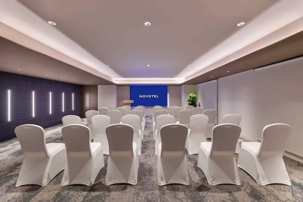 Ein Konferenzraum mit Reihen weißer Stühle, einem Rednerpult und einer blauen Wand mit der Aufschrift 'NOVOTEL'.