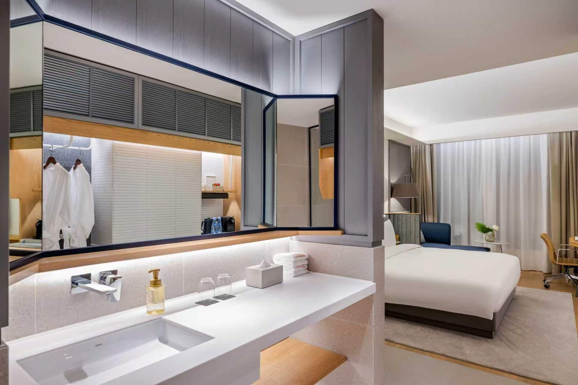 A modern hotel room with a large mirror, a white sink, a bed, and a bedside table.