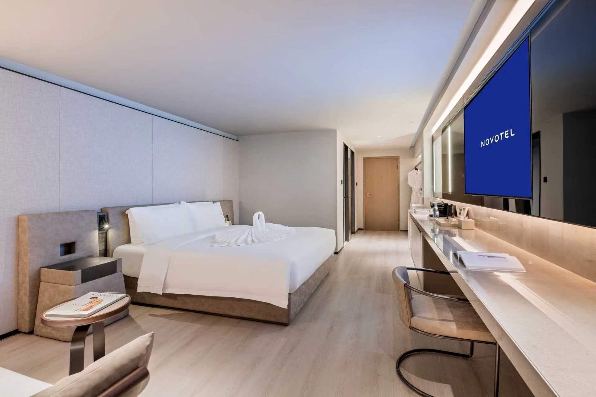 A modern hotel room with a bed, bedside table, chair, and a large mirror with a TV.