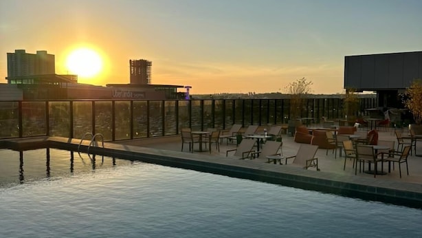 A swimming pool with lounge chairs and tables, a sunset in the background, and a cityscape. - Novotel Uberlândia (Uberlandia)