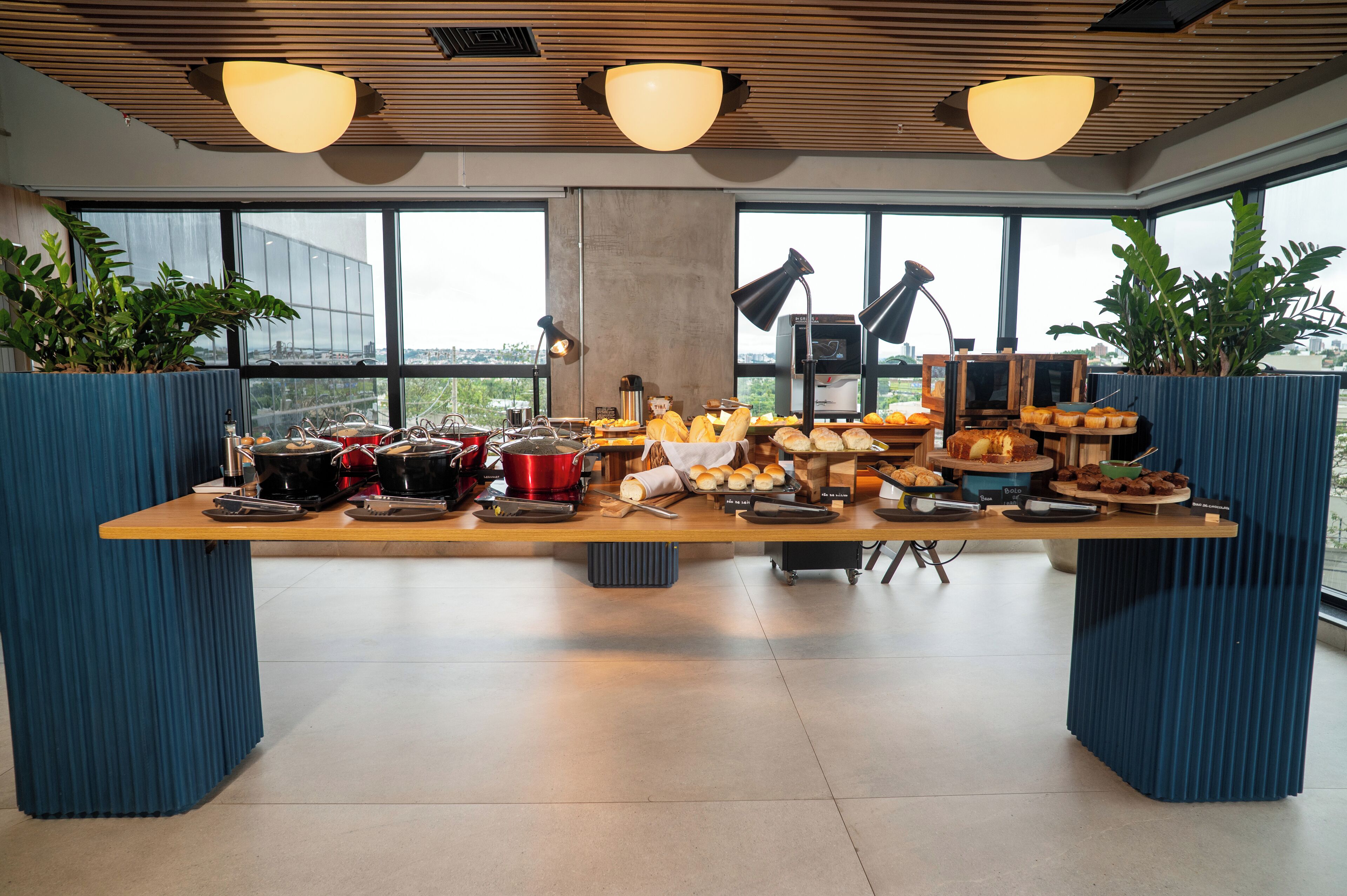 A long table with various food items and kitchen appliances, set in a modern room with large windows and blue planters.