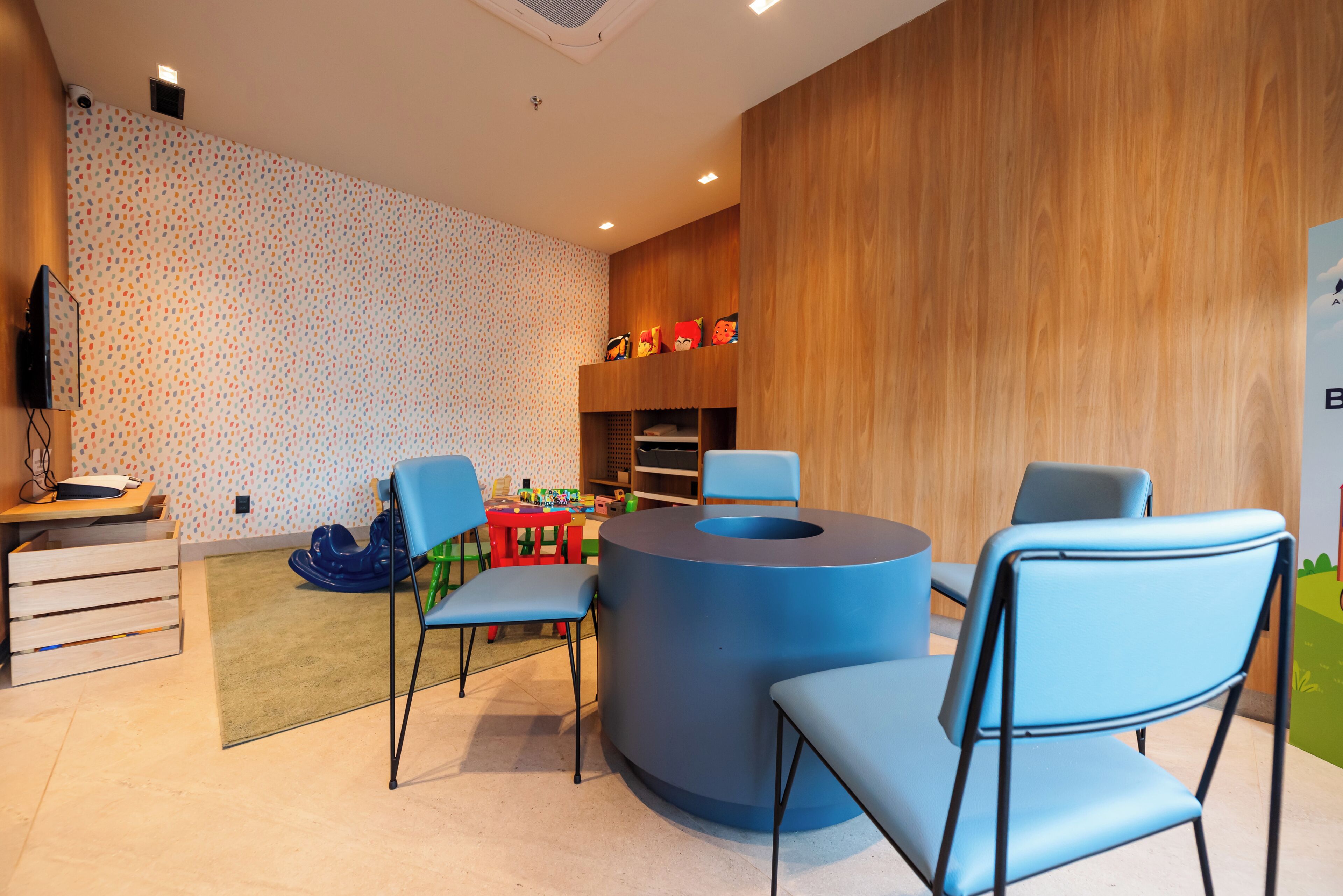 Uma área de recreação infantil com uma mesa azul circular, cadeiras azuis e brinquedos coloridos.